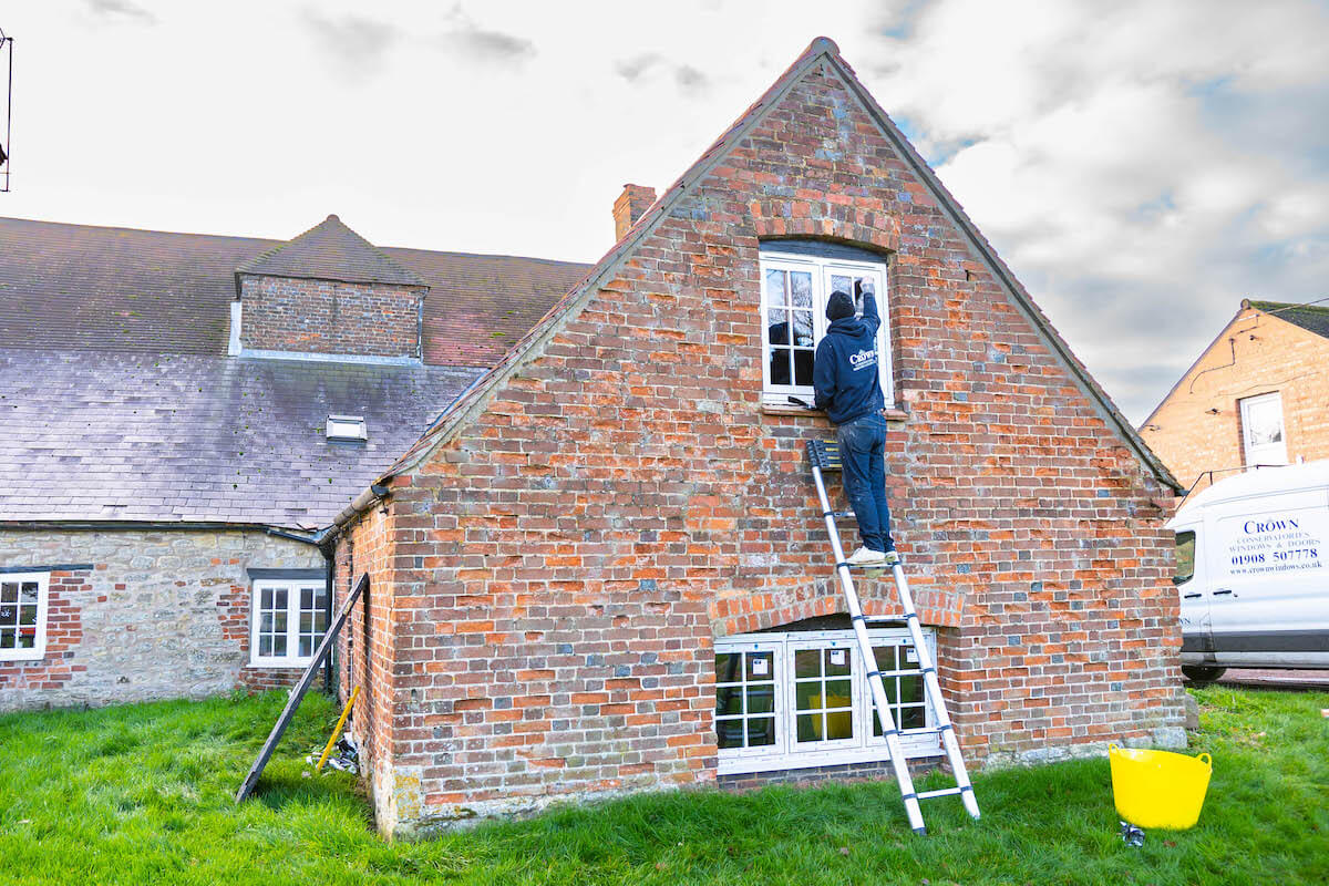 Crown Windows installer replacing white Upvc flush window on a brick cottage using a ladder.